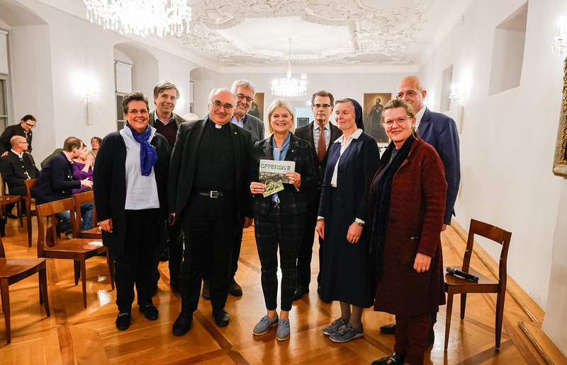 Verteidigungsministerin Klaudia Tanner mit der letzten Ausgabe der 'Offensive' der Katholischen Aktion zum Thema 'Frieden', und Bischof Wilhelm Krautwaschl, umringt von Vertreter:innen der einzelnen Kurien des KLRÖ